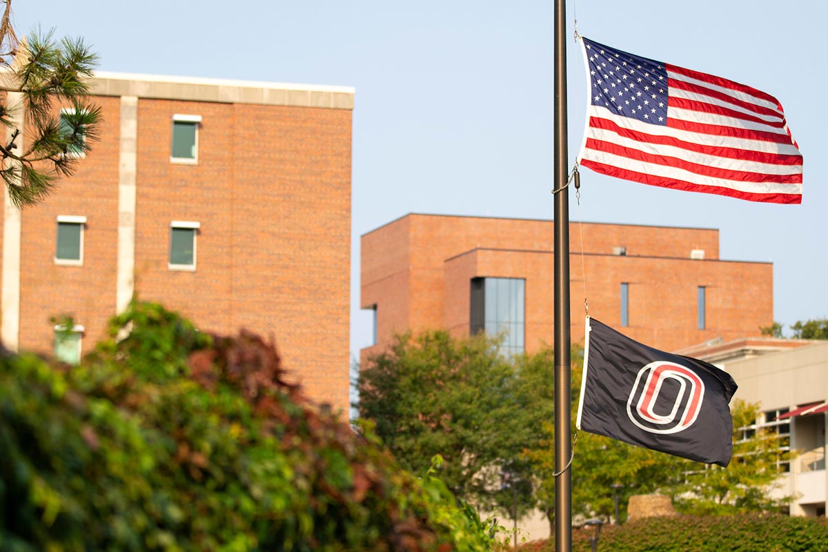 A photo of the UNO flag, lowered, next to the Pep Bowl on Dodge Campus