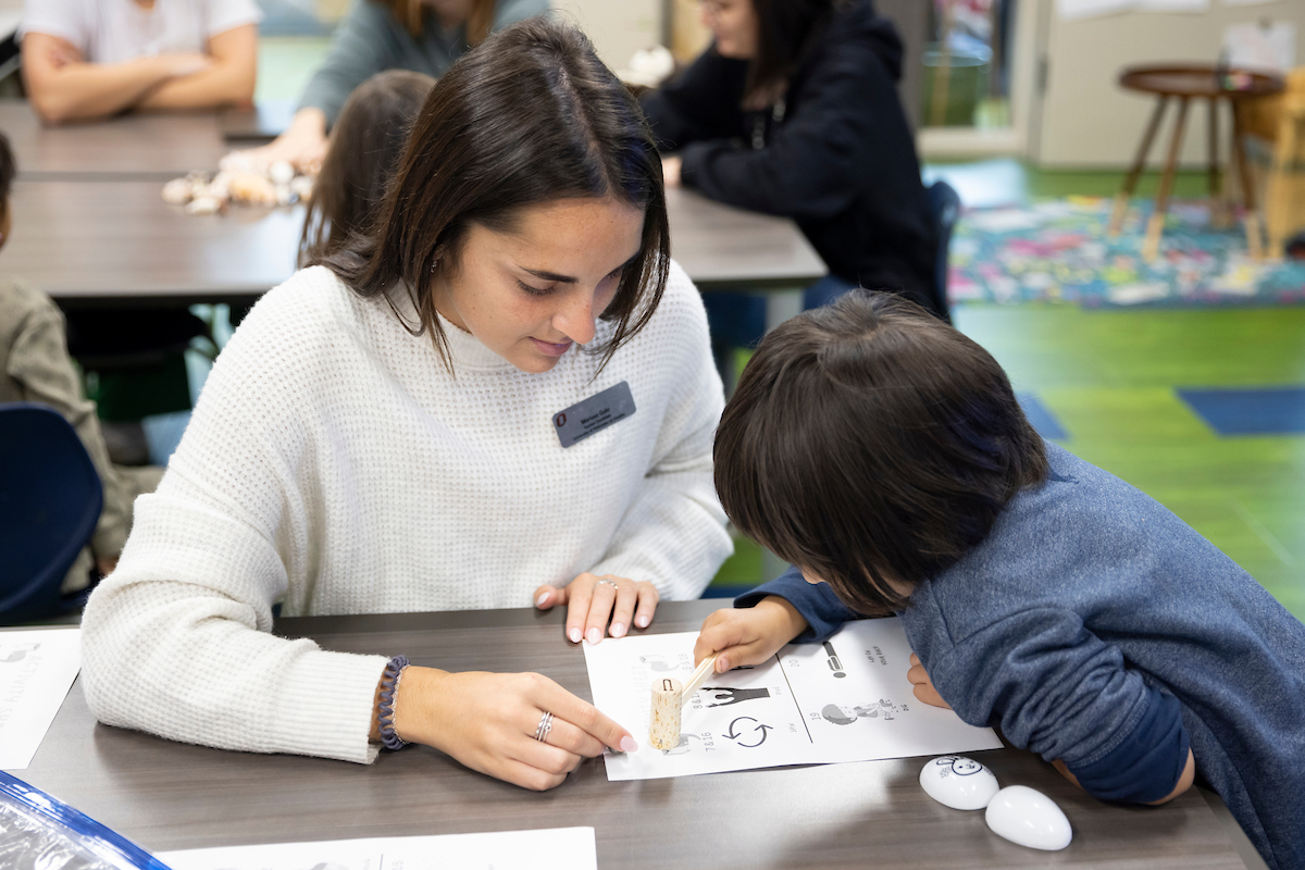 A young adult works with a preschooler on an assignment in a clasroom