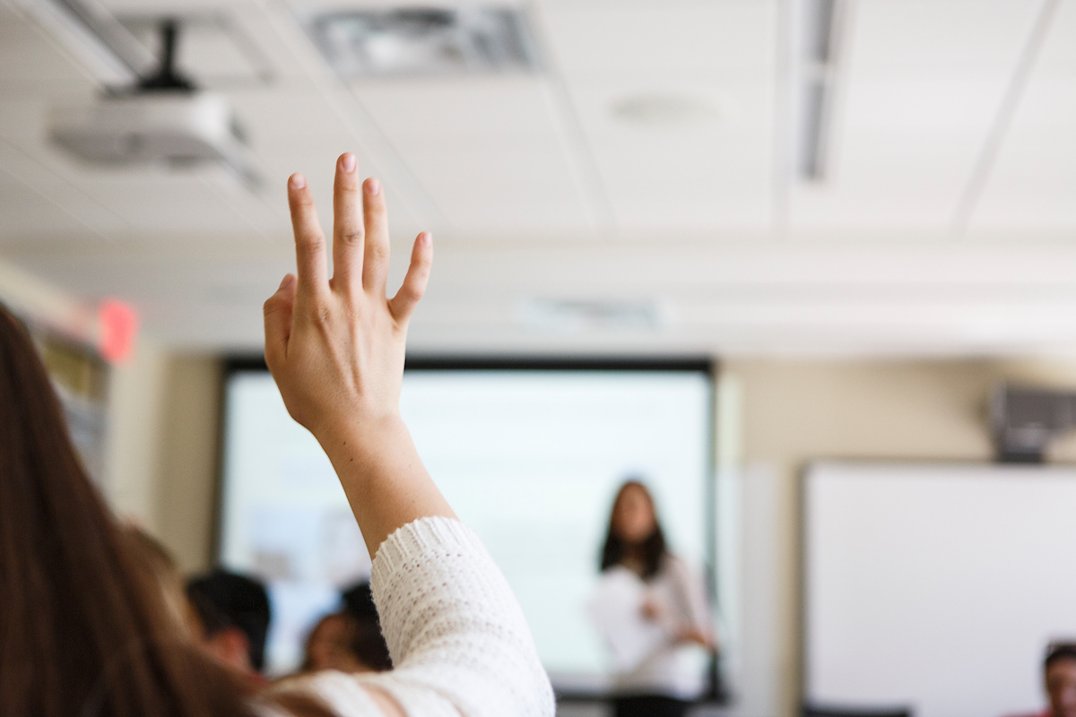 Image of a hand being raised in a classroom, background is blurred/not in focus.