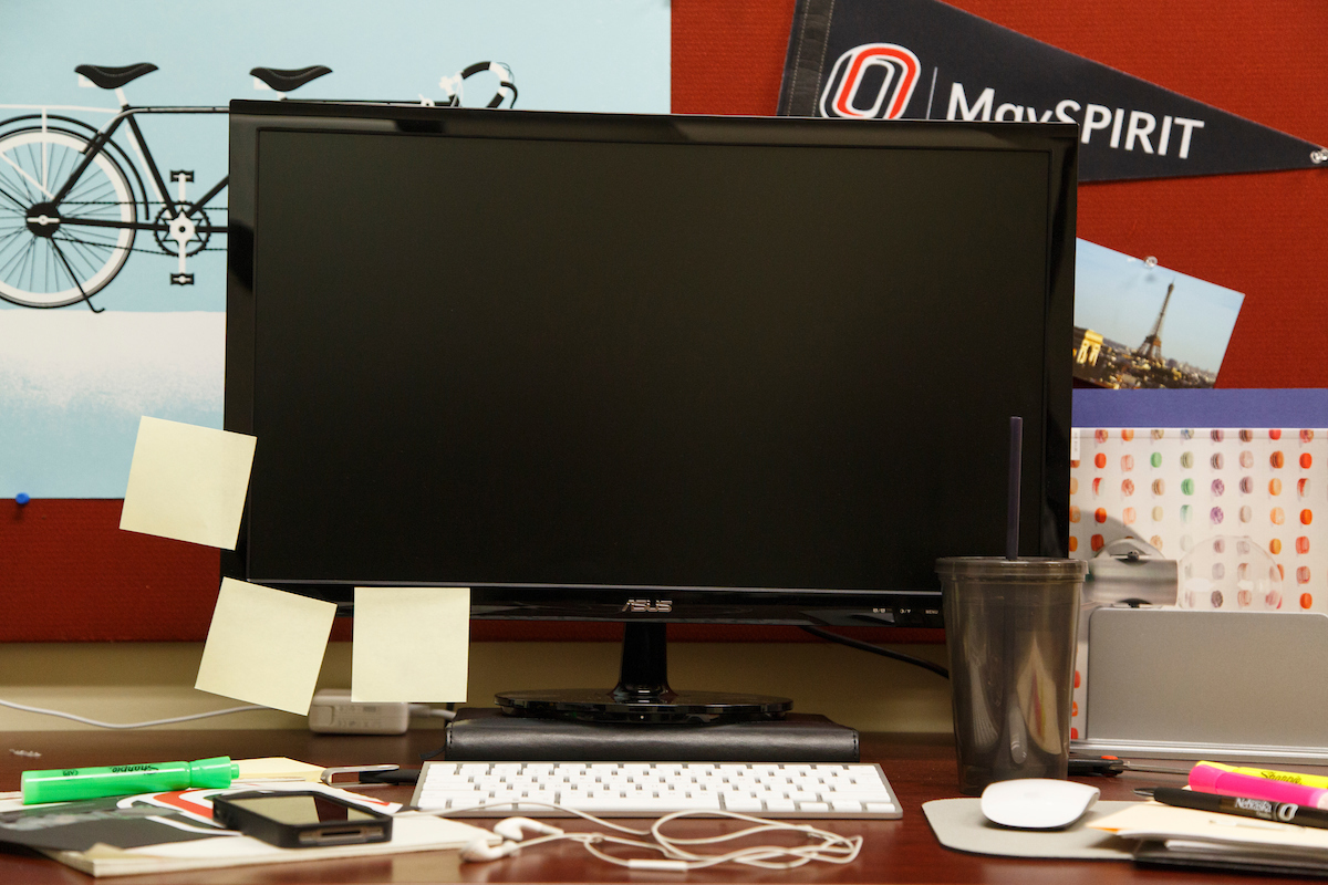 Computer desk with a monitor, cluttered with student-related items such as post-it notes, ear buds, keyboard, and writing utensils.