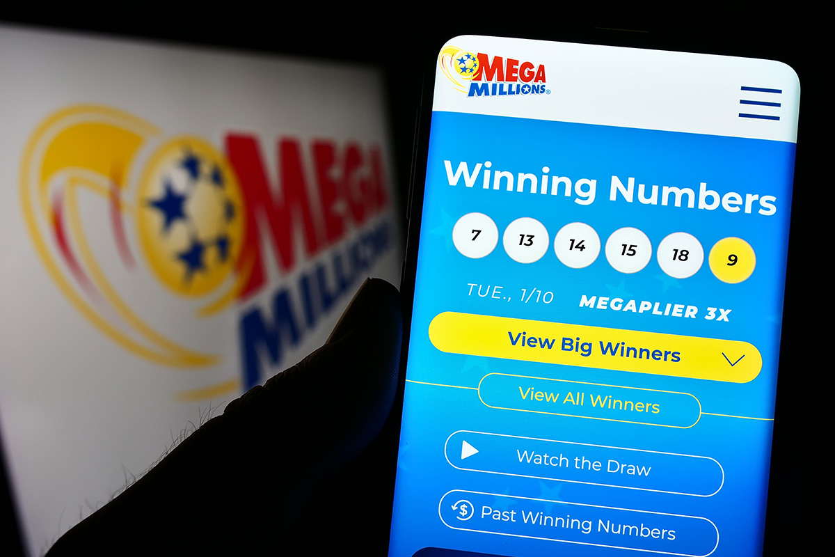 A person holding a cell phone with the Mega Millions winning numbers displayed. Mega Millions also appears on a TV in the background.