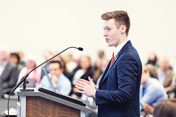 Student speaks at a podium to an audience in a formal event setting.