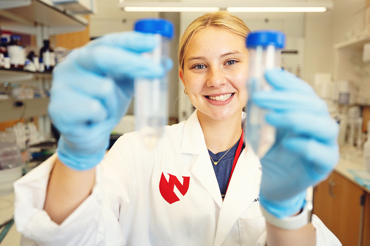 A student in a lab coat holds up two test tubes in a science lab.