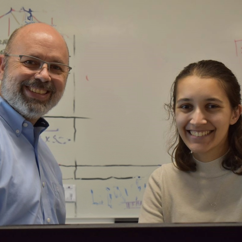 Professor and student stand at a computer in a classroom with a white board behind them