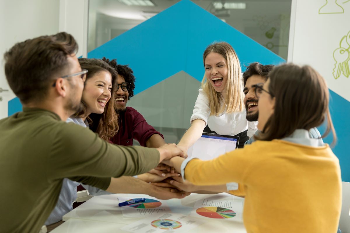 A group of young professionals gather around a table, laughing and stacking their hands together in a gesture of teamwork. Charts, graphs, and a laptop are visible on the table, indicating a collaborative project or brainstorming session.