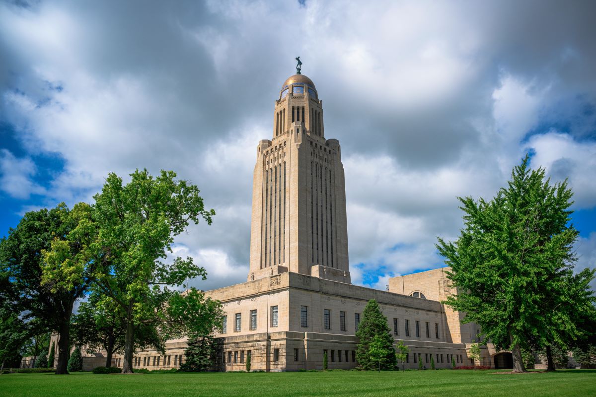 The Nebraska State Capitol building stands tall against a partly cloudy blue sky. Its distinctive Art Deco tower, topped with a golden dome and the "Sower" statue, is surrounded by lush green trees and a well-manicured lawn.
