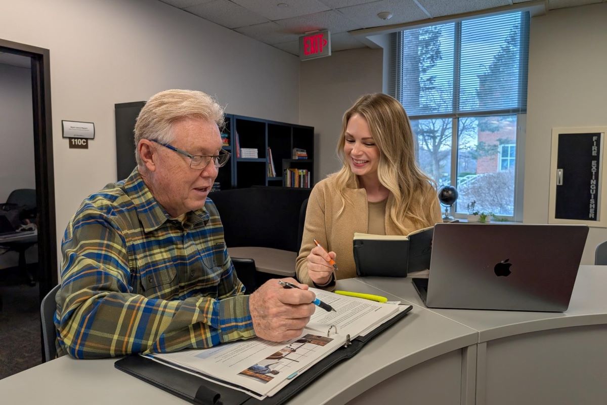 An older man in glasses and a plaid shirt discusses a document with a younger woman in a beige sweater. They sit at a table with a laptop and papers in an office setting.