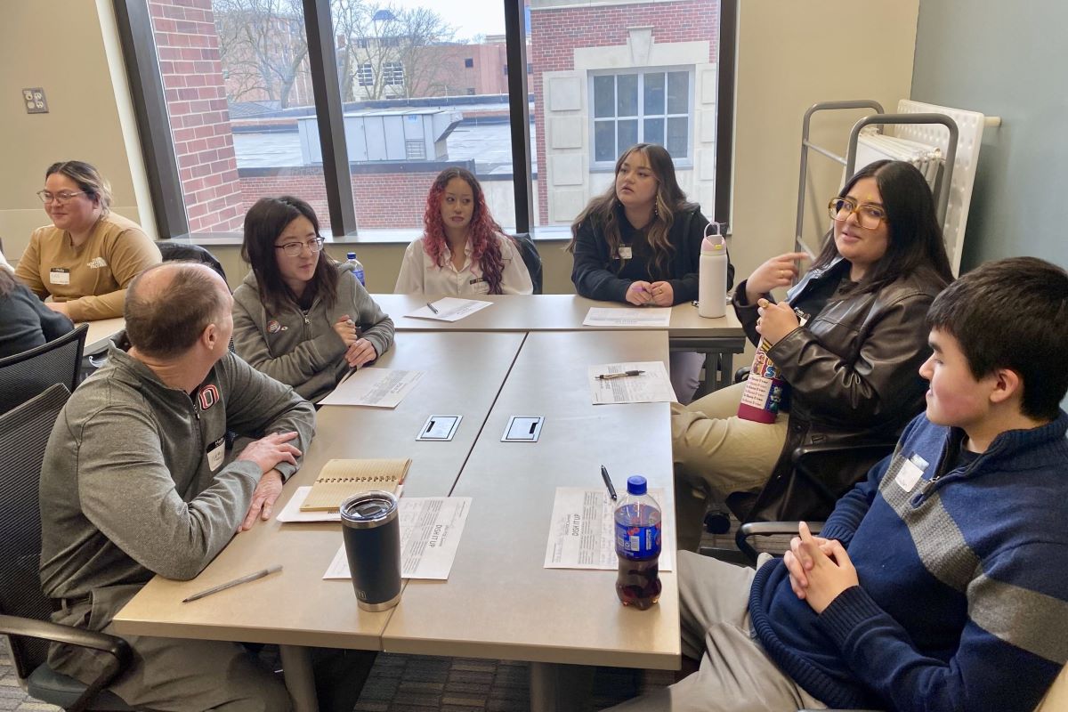 A group of students and faculty sit around a table in a classroom, engaged in discussion. Papers, notebooks, and drinks are scattered across the table.