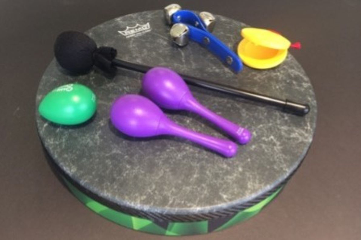 A collection of colorful percussion instruments displayed on a round, marble-patterned drum. The set includes a green egg shaker, two purple maracas, a black-handled mallet with a foam head, a blue wrist jingle bell strap, and a pair of yellow castanets.