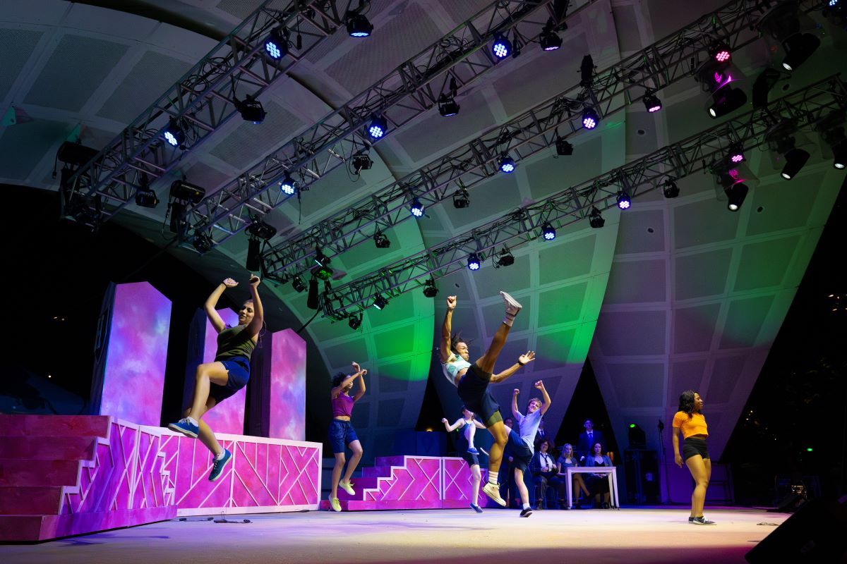 Dancers perform an energetic routine on a well-lit stage with pink and green lighting. They leap into the air, arms raised, against a backdrop of geometric stage props.
