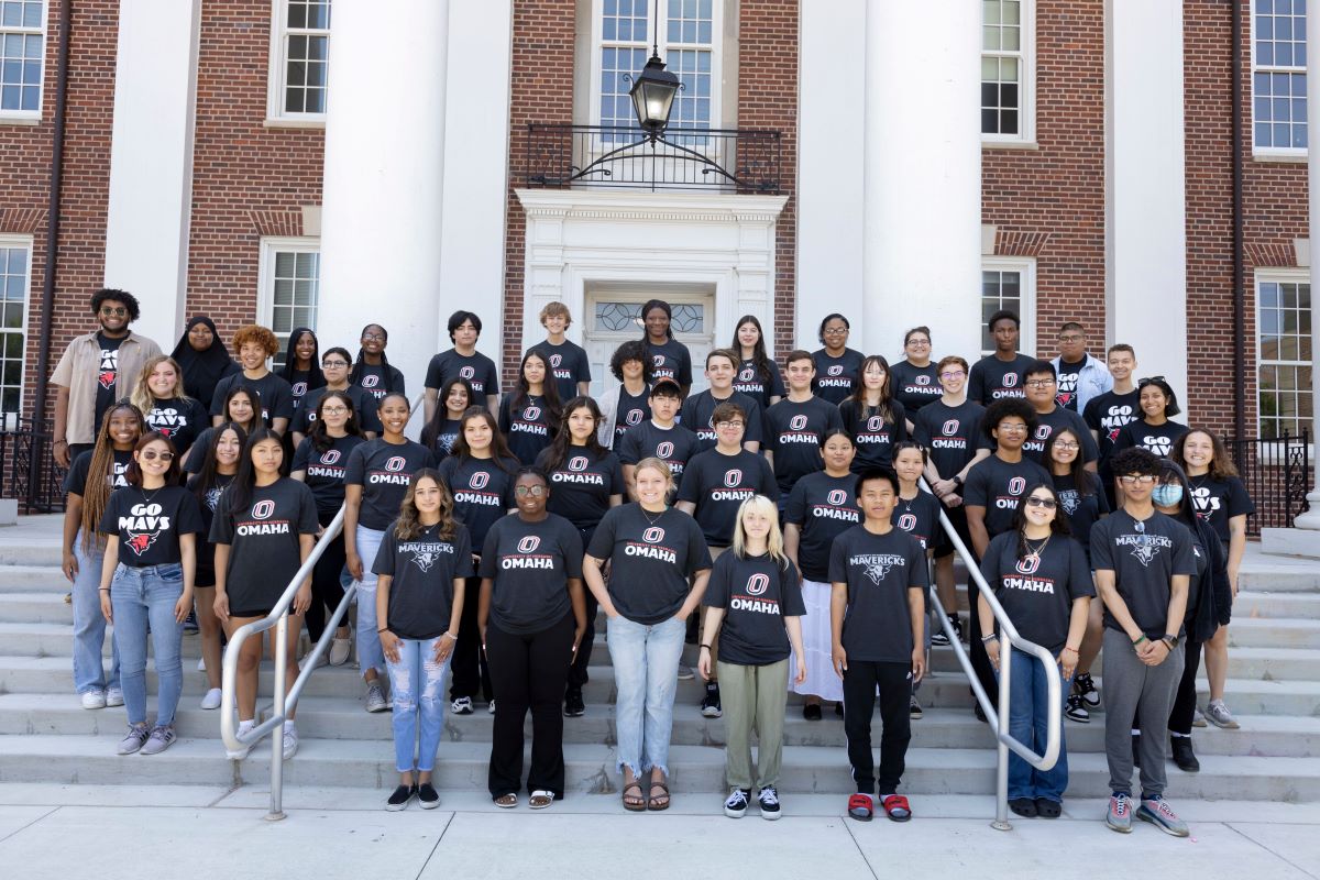 A group of students in matching black "UNO Omaha" T-shirts pose on the steps of a red brick building with large white columns. The students smile, standing in multiple rows.