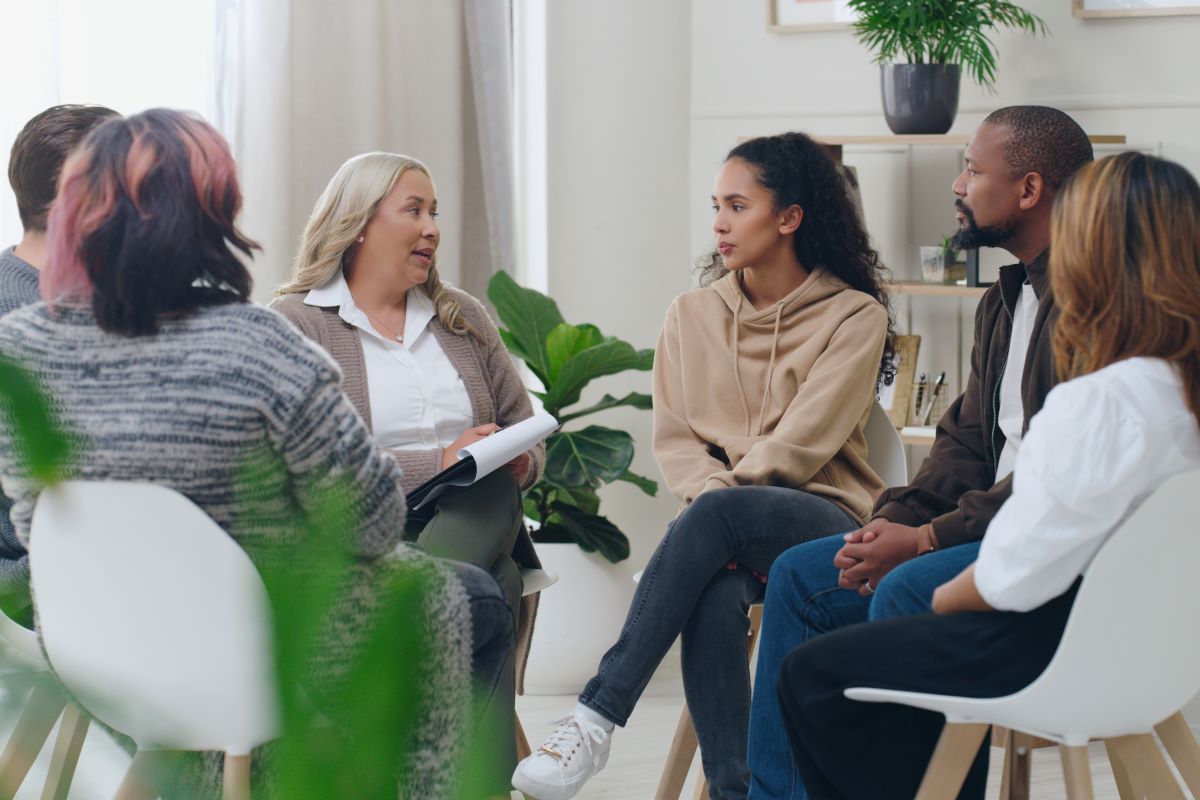 A diverse group of adults seated in a circle during a group discussion or coaching session, with a woman holding a notebook speaking to the group.