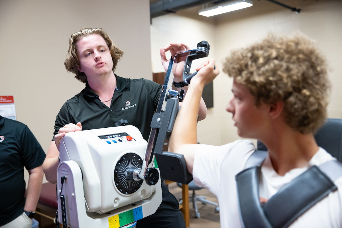 A student in a UNO Biomechanics shirt adjusts a large piece of exercise testing equipment while another student, seated and strapped in, performs an arm movement test.