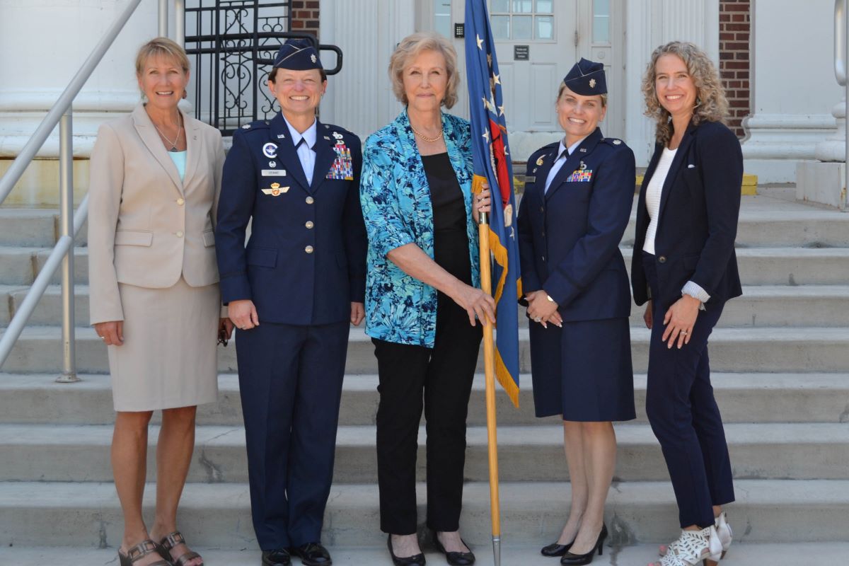 Five women, including military personnel, pose with a flag in front of a building’s steps.