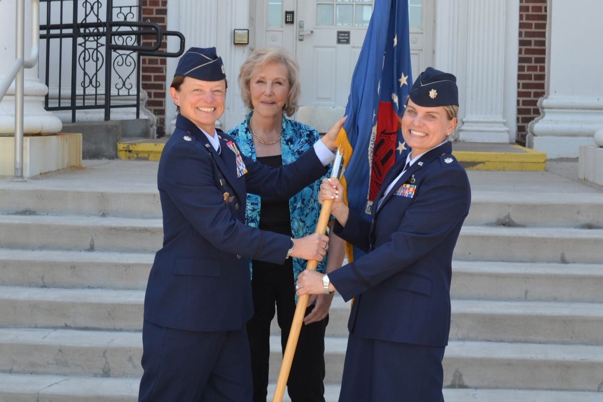 Two uniformed officers smile while passing a ceremonial flag, with a woman standing behind them.