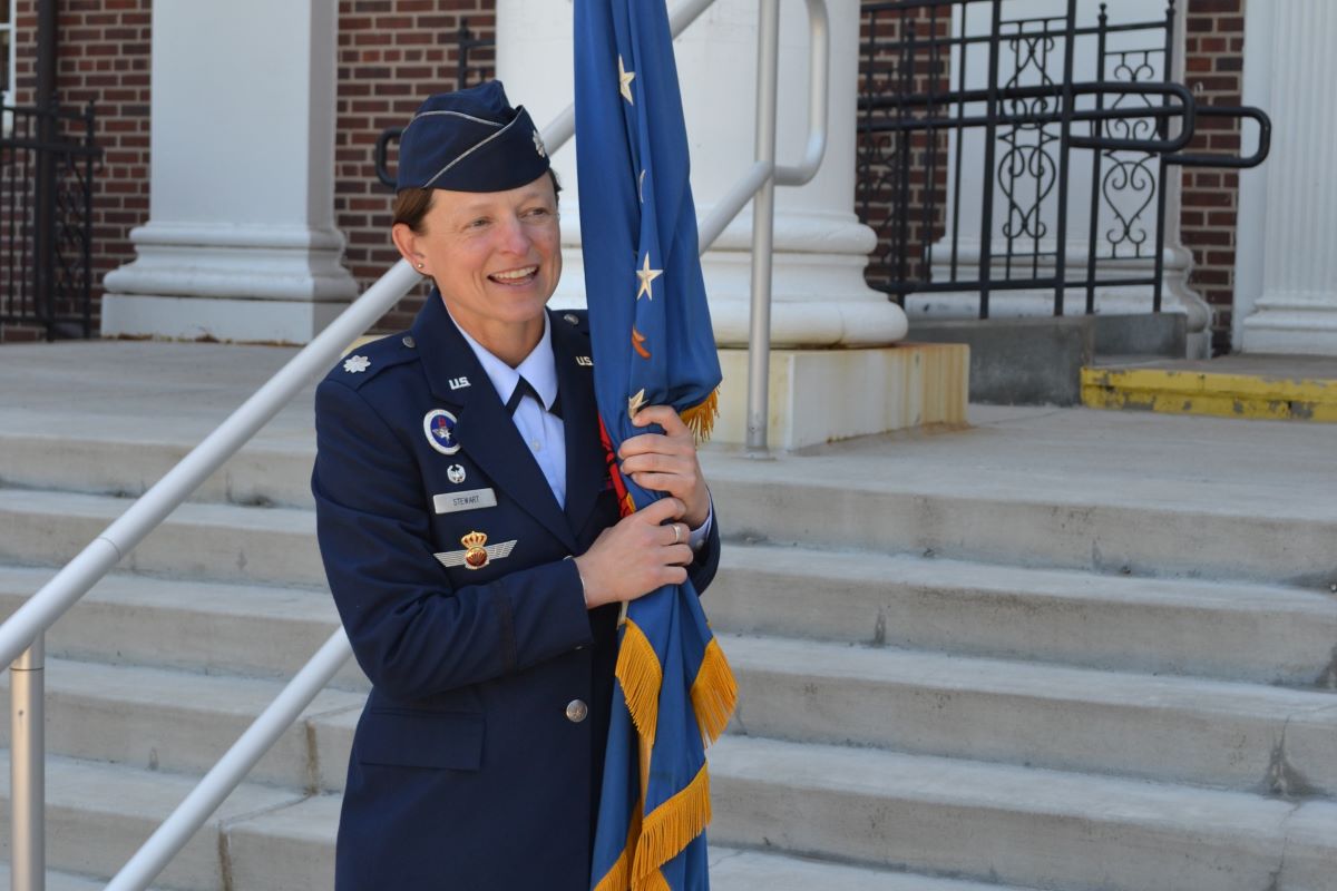 Lt. Col. Stewart stands on building steps smiling and holding a ceremonial flag.