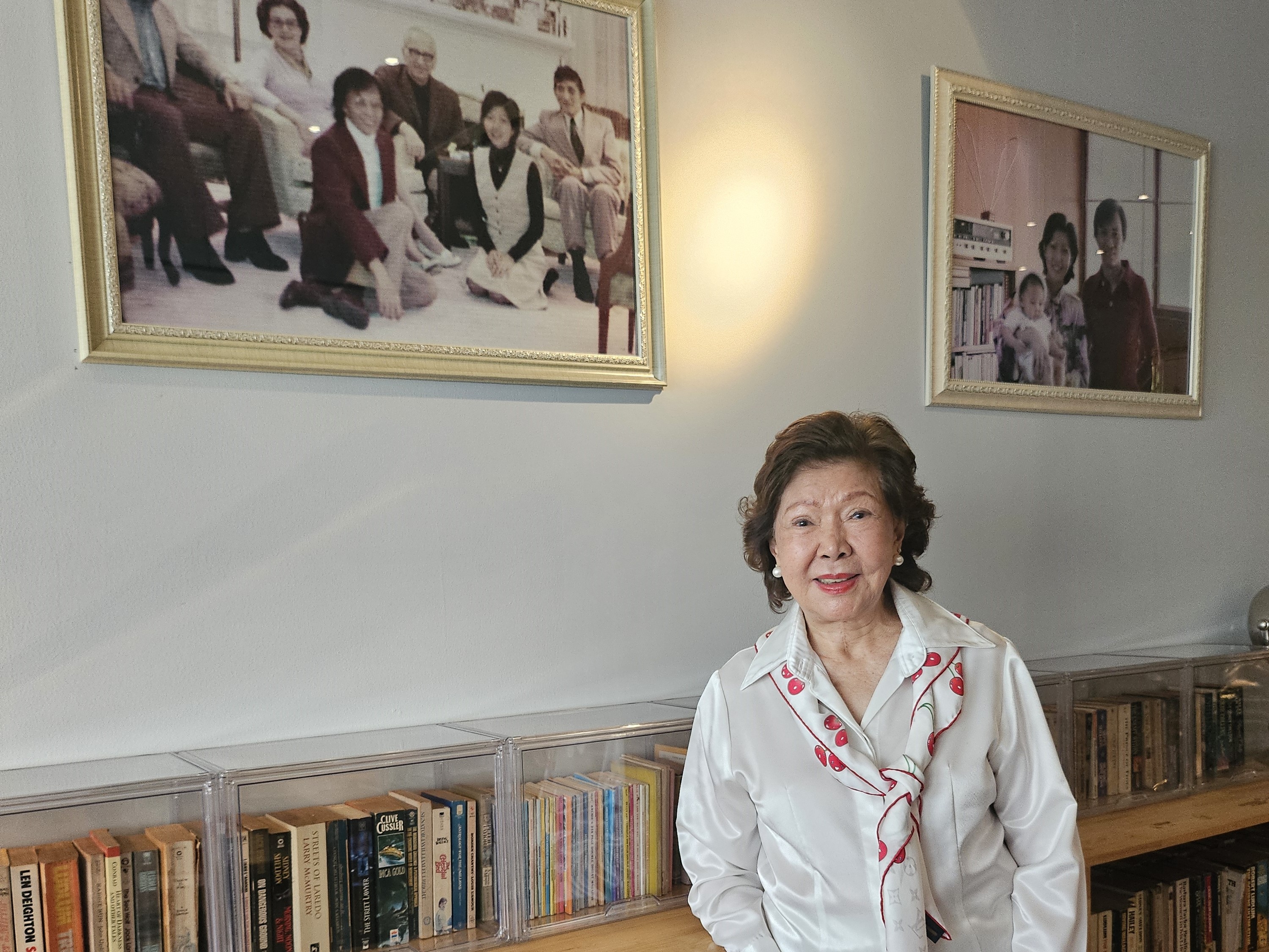 An older woman smiling while standing in front of a bookshelf filled with books. Framed family photographs hang on the wall behind her.