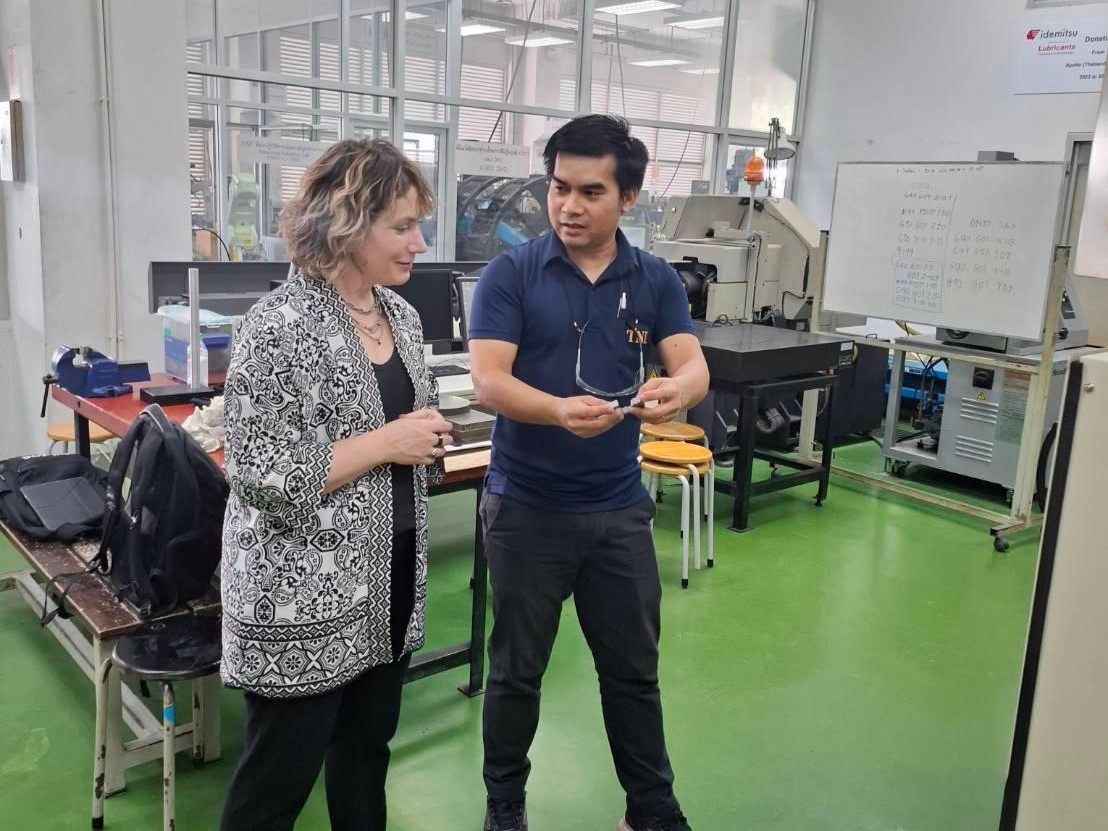 A man demonstrating a small object to a woman inside a lab or workshop space, with machinery and equipment visible in the background.