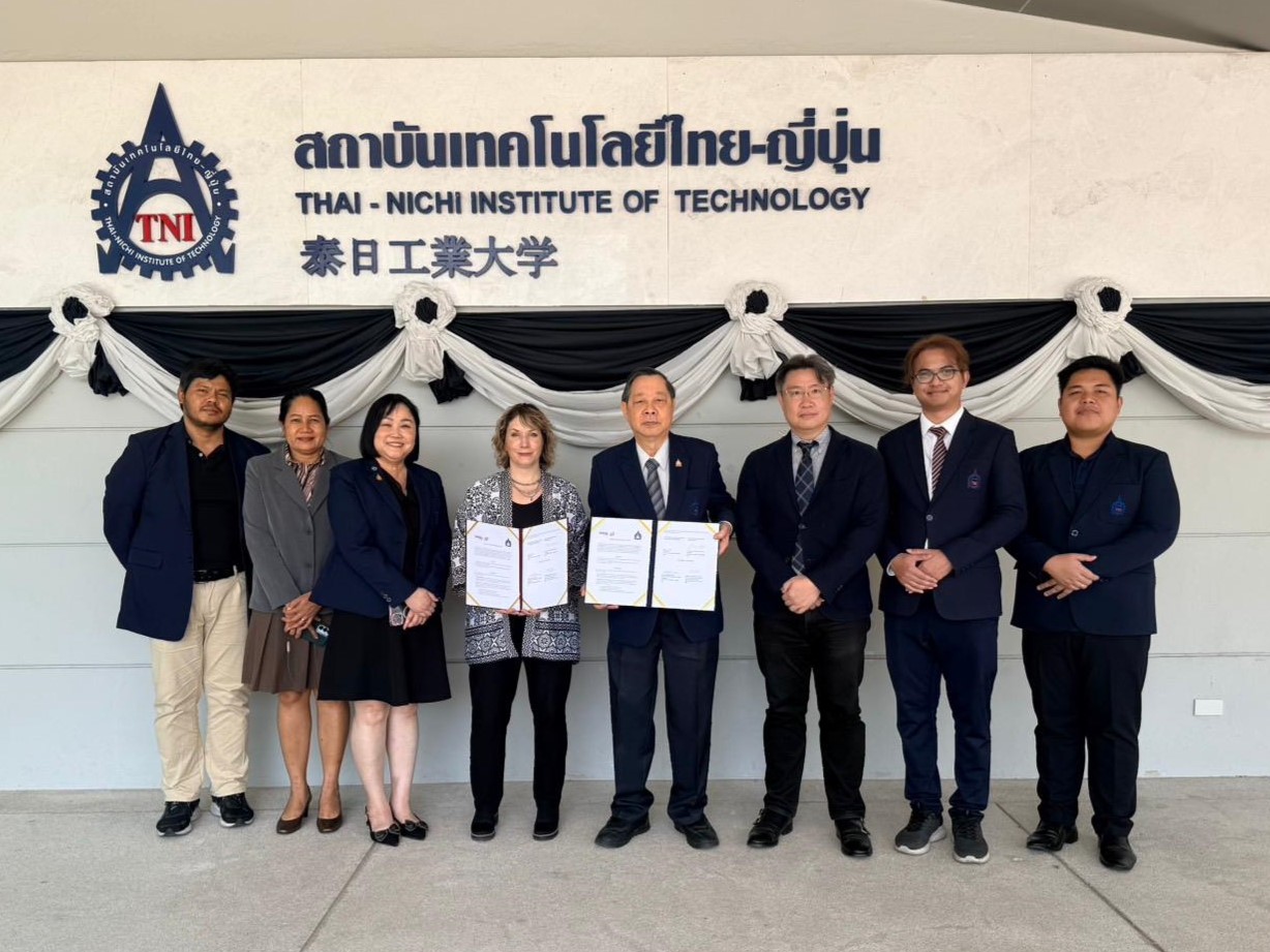A group of nine people standing in front of a sign for the Thai-Nichi Institute of Technology. Two individuals in the center hold open documents during a formal signing photo.