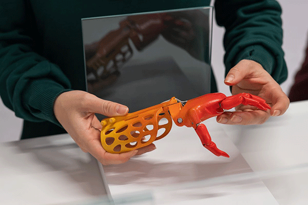 A person wearing a green sweater holds a 3D-printed prosthetic arm with an orange and red design. A reflective panel behind the arm shows its mirrored image.