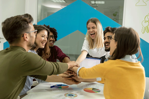 A group of young professionals gather around a table, laughing and stacking their hands together in a gesture of teamwork. Charts, graphs, and a laptop are visible on the table, indicating a collaborative project or brainstorming session.