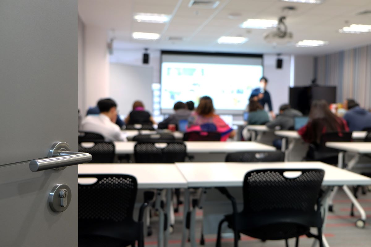 View from an open classroom door showing students seated at tables and listening to a presenter at the front of the room.