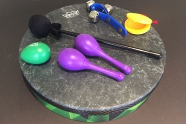 A collection of colorful percussion instruments displayed on a round, marble-patterned drum. The set includes a green egg shaker, two purple maracas, a black-handled mallet with a foam head, a blue wrist jingle bell strap, and a pair of yellow castanets.