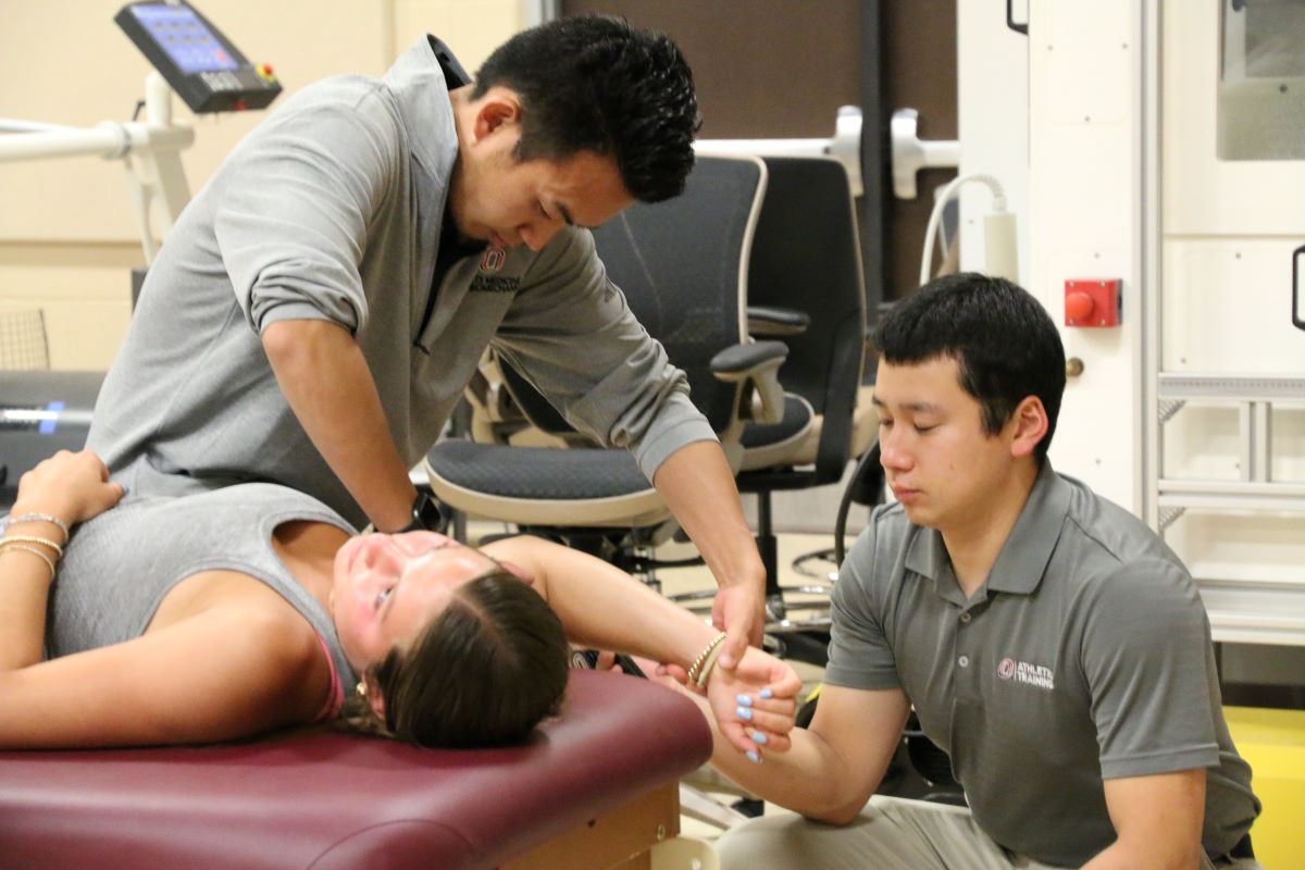Two male students in UNO Athletic Training uniforms assist a female student lying on a treatment table by mobilizing her shoulder and arm in a classroom lab setting.