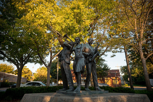 A bronze statue of three jazz musicians—one playing a trombone, another singing, and the third playing a saxophone—stands under lush green trees. The statue captures the energy of North Omaha’s rich jazz history, with warm sunlight filtering through the branches.