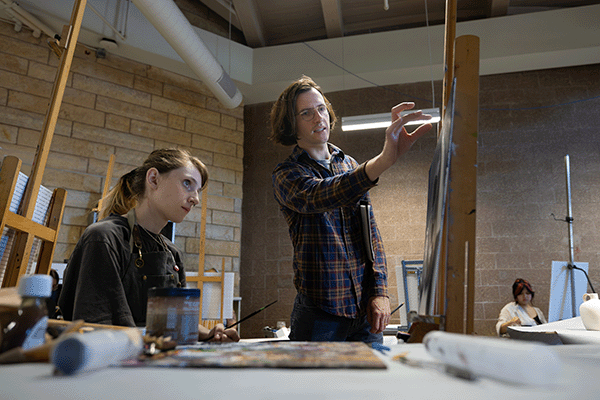 a man points at a student's easel as the woman student looks on
