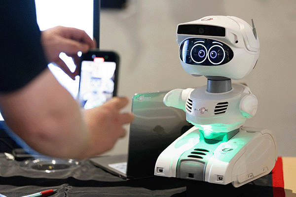 a person holds a mobile phone in the foreground to take a picture of a small robot on a table.