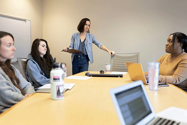 a woman stands with a clipboard listening to students at a table