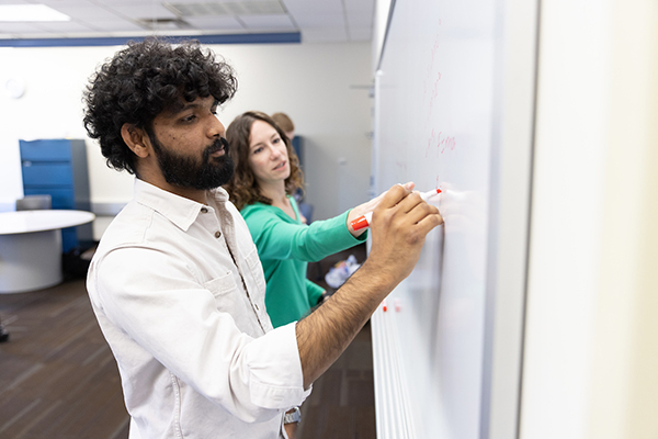 a male student writes on a whiteboard as a female professor looks on
