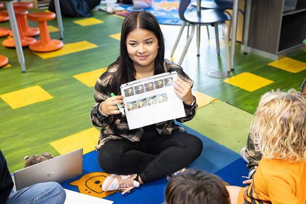 A female student sitting in a classroom reading a book to children.