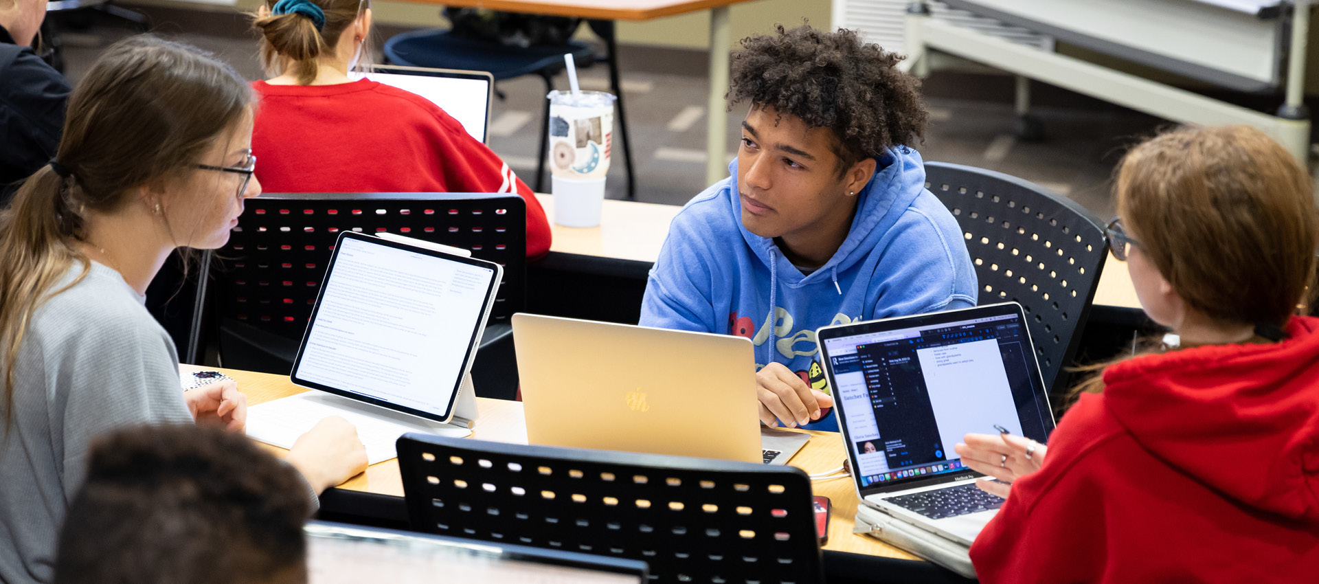 Students sitting in a classroom at a round table with their laptops open having a discussion.