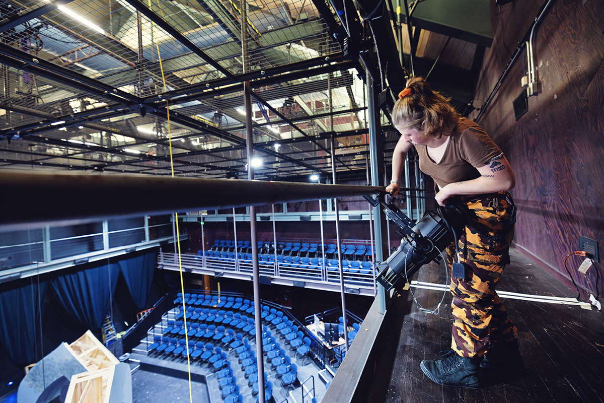 A student works with lighting equipment above a theater stage, preparing for a production.