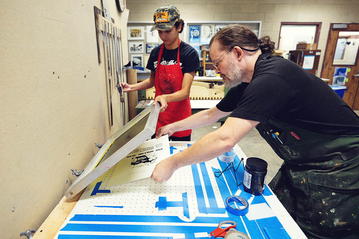 An instructor guides a student through a screen-printing process in an art studio.