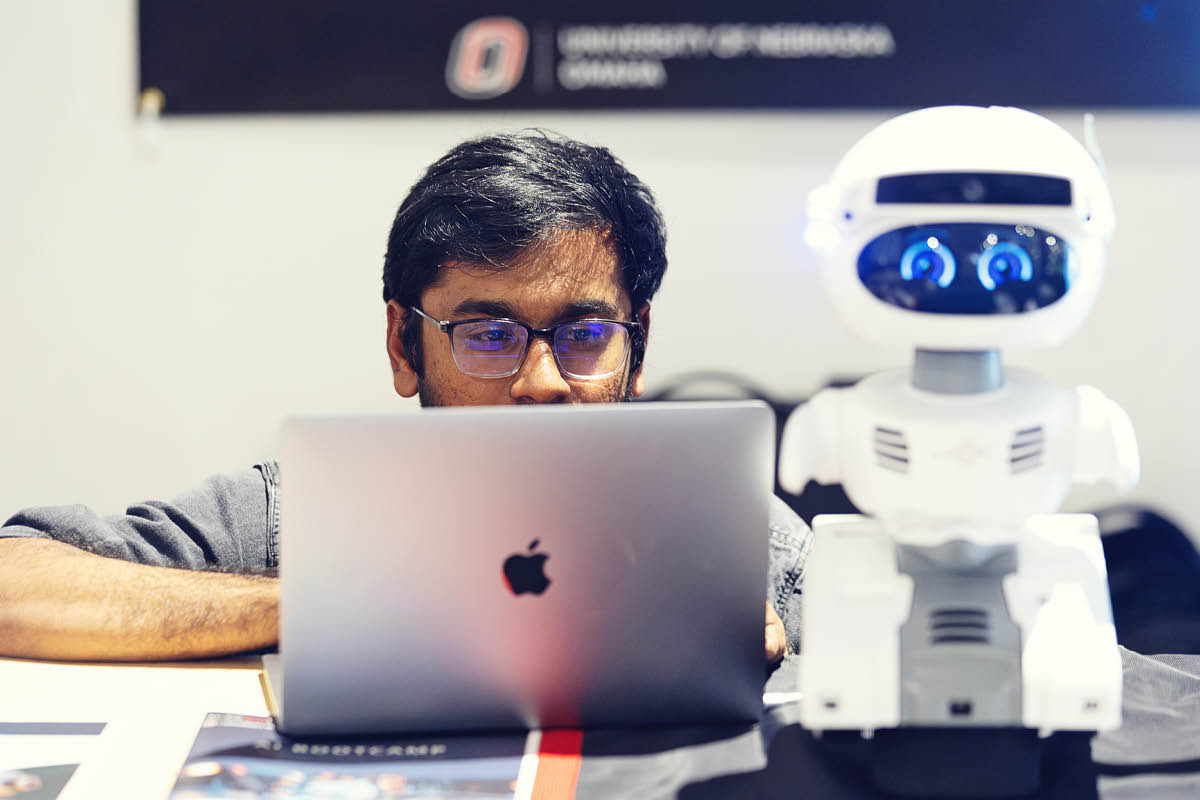 A student works on a laptop beside a small robot during a technology or robotics project.