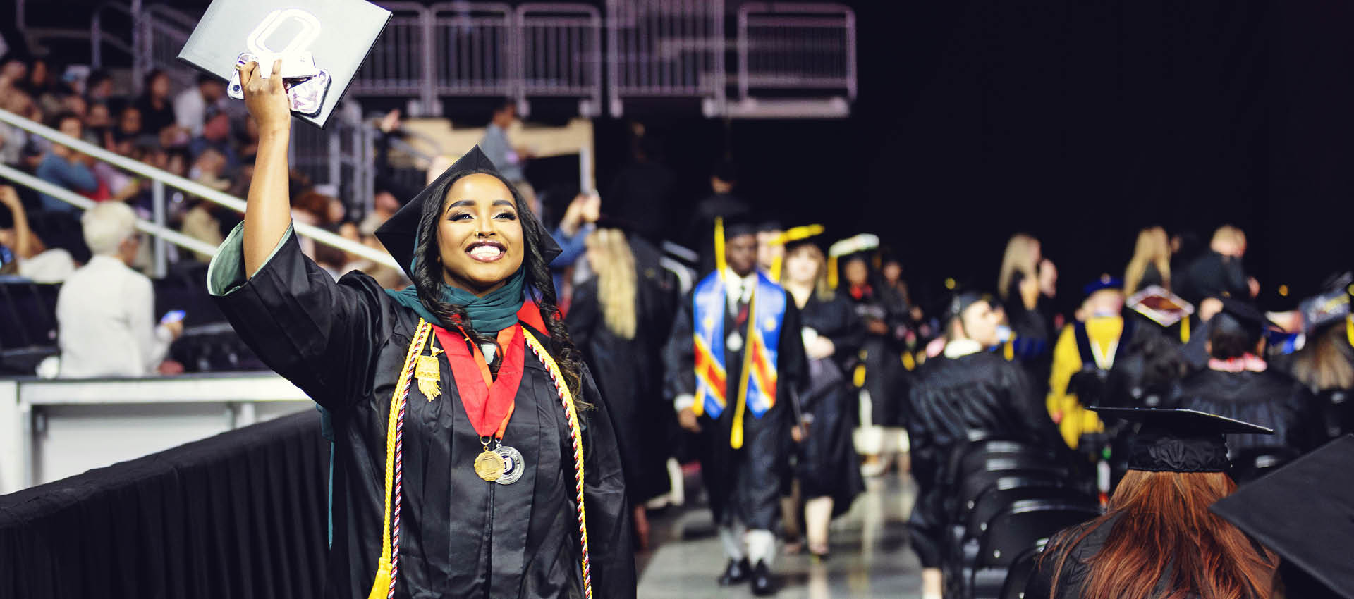 Female graduate is walking down the aisle at commencement holding her diploma up in the air smiling.