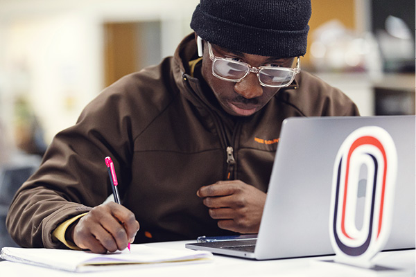 male student works on laptop while taking notes in a notebook