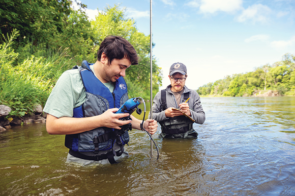 Two students stand in a river collecting water samples as part of a research project.