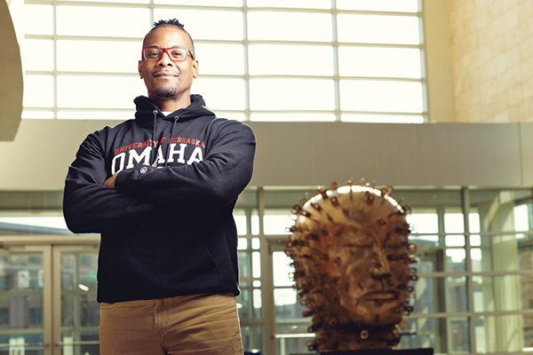 A student wearing an Omaha sweatshirt smiles with arms crossed inside a campus building.