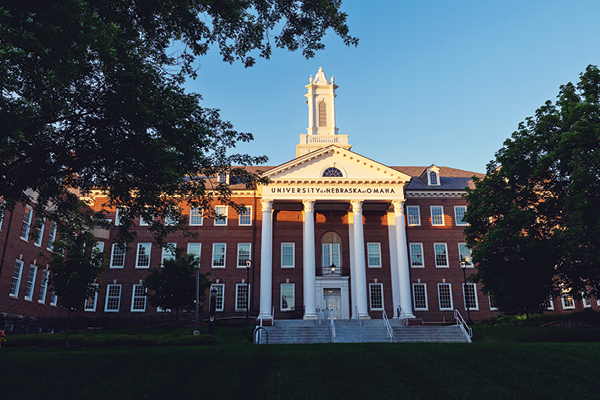 The UNO Arts and Sciences Hall building stands under a clear blue sky with trees framing the view.