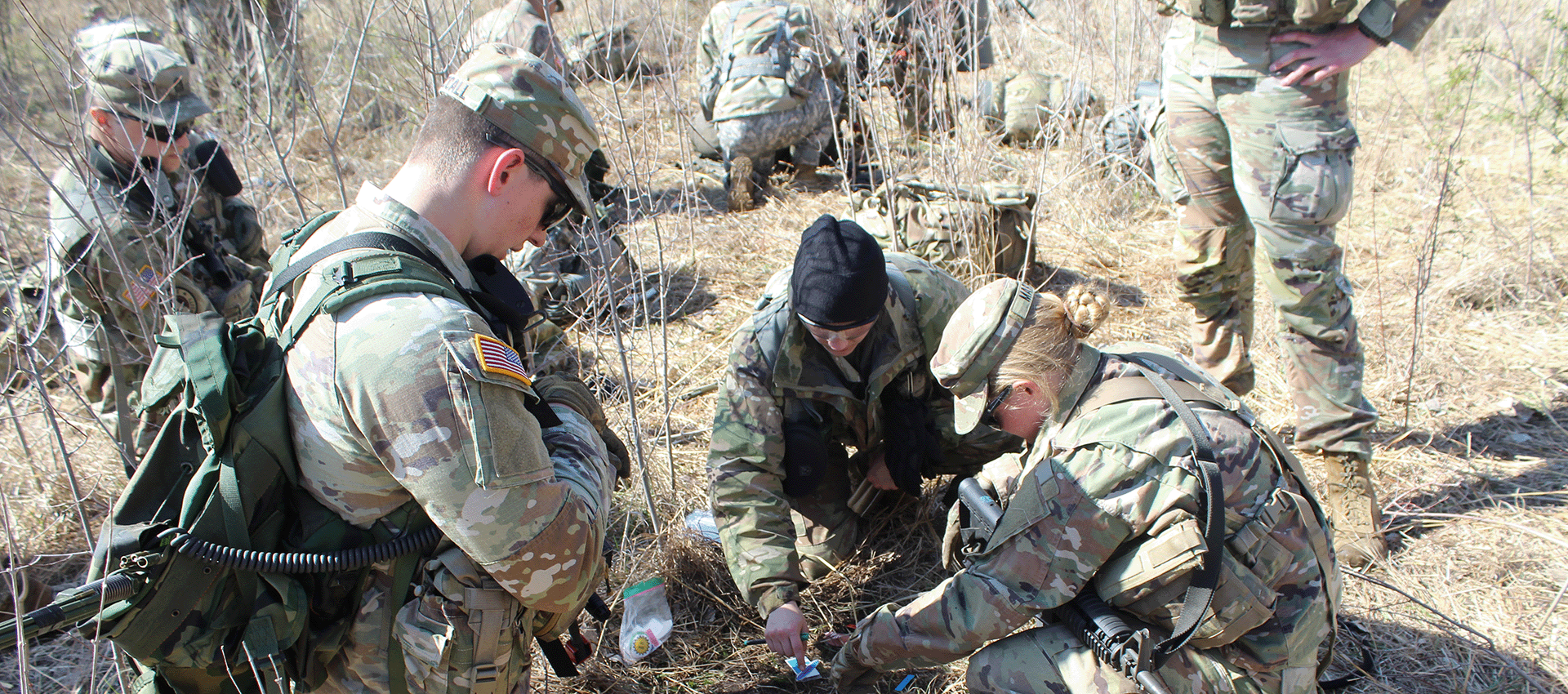 Soldiers in camouflage gear work together on the ground in a dry, brushy field during a training exercise.