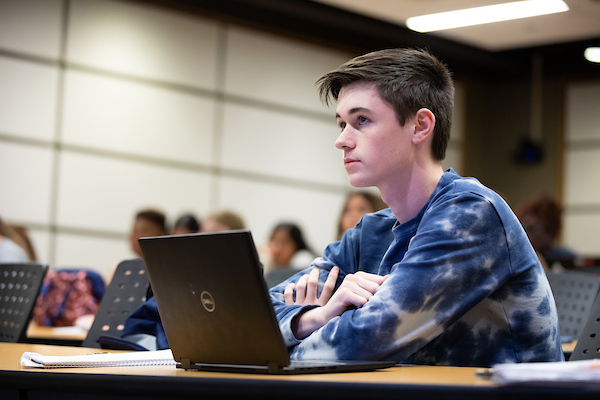 a young male student sits in class with his laptop in front of him on a table.