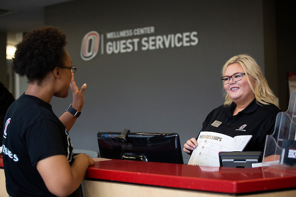 a woman shows a sign up pamphlet to another woman as they stand at a desk.
