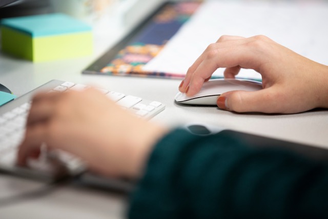 Close-up of a person using a computer workstation, with one hand on a mouse and the other near a keyboard. Colorful sticky notes and office materials are visible on the desk in the background.