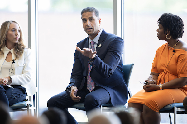 Three panelists participate in a discussion at a Future of Work event. The speaker in the center, dressed in a navy suit, gestures while addressing the audience, as two fellow panelists listen attentively. The discussion takes place in a bright conference space with large windows in the background.