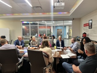 A large lecture hall filled with attendees seated at long desks, listening to a speaker presenting at the front of the room beside a large projection screen.