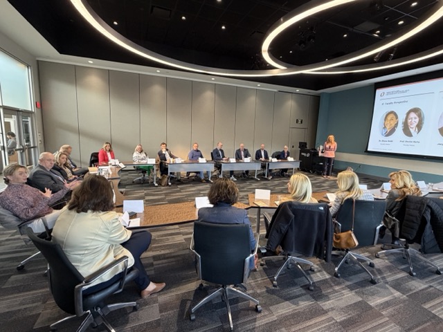 A large lecture hall filled with attendees seated at long desks, listening to a speaker presenting at the front of the room beside a large projection screen.
